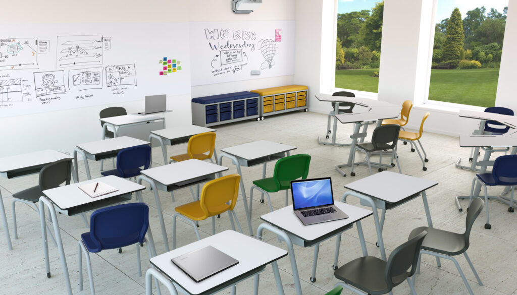 Un aula moderna con Mobiliario Escolar de colores, pupitres blancos, ordenadores portátiles y cuadernos. Los grandes ventanales dejan ver la vegetación del exterior, mientras las pizarras blancas muestran bocetos y apuntes. La sala está bien iluminada y vacía de alumnos.