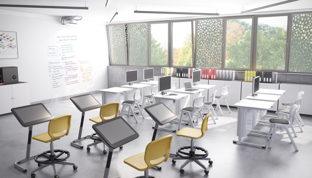 Un aula moderna con grandes ventanales, luz solar, pupitres blancos, sillas Mobiliario Escolar amarillas y blancas, monitores de ordenador, pupitres móviles con tabletas negras y una pizarra blanca cubierta de notas de colores. En el exterior se ven árboles.